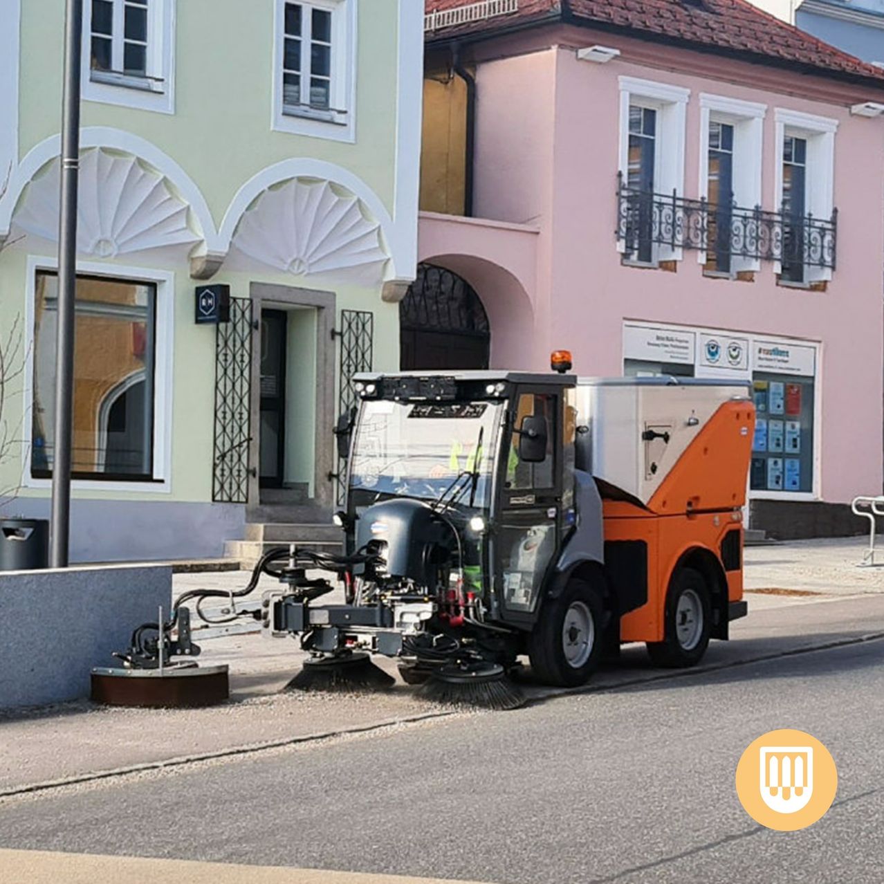 Ein Straßenreinigungsfahrzeug ist am Straßenrand vor einem Gebäude mit dekorativen weißen Bögen und Fenstern geparkt. Das Fahrzeug ist orange und weiß und hat eine Bürste daran befestigt.