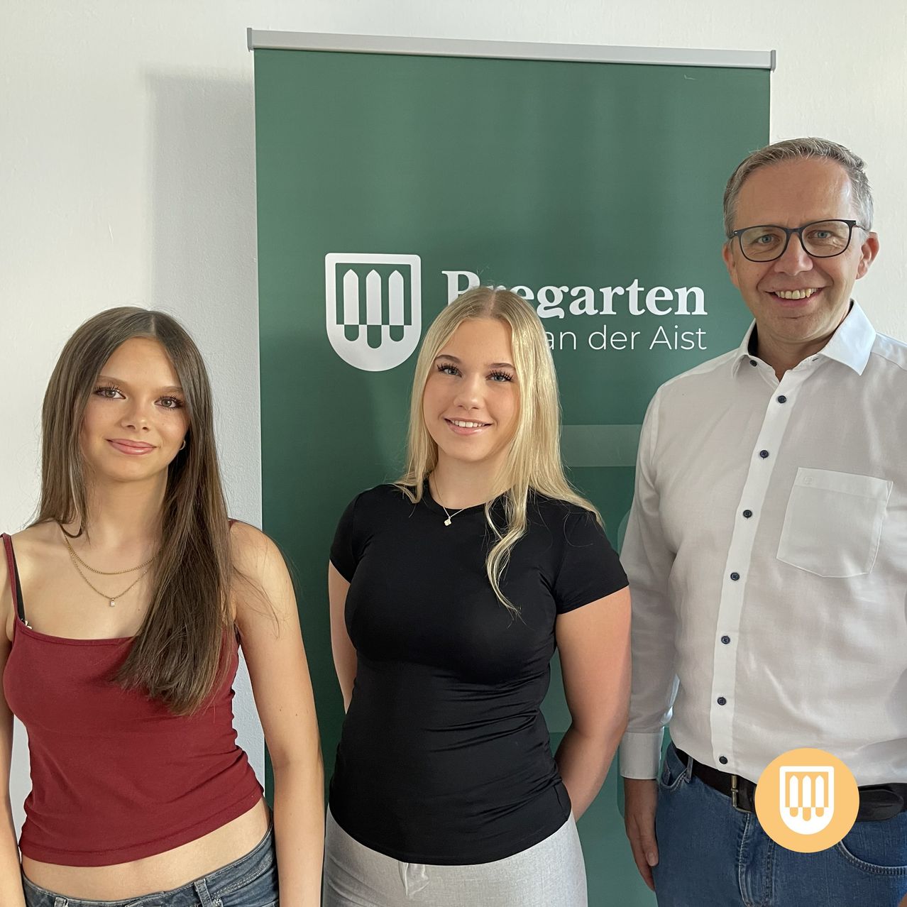 Three individuals stand in front of a green banner with the text 'Regarten an der Aist'. The woman on the left wears a maroon tank top, the woman in the middle a black t-shirt, and the man on the right a white shirt and glasses.
