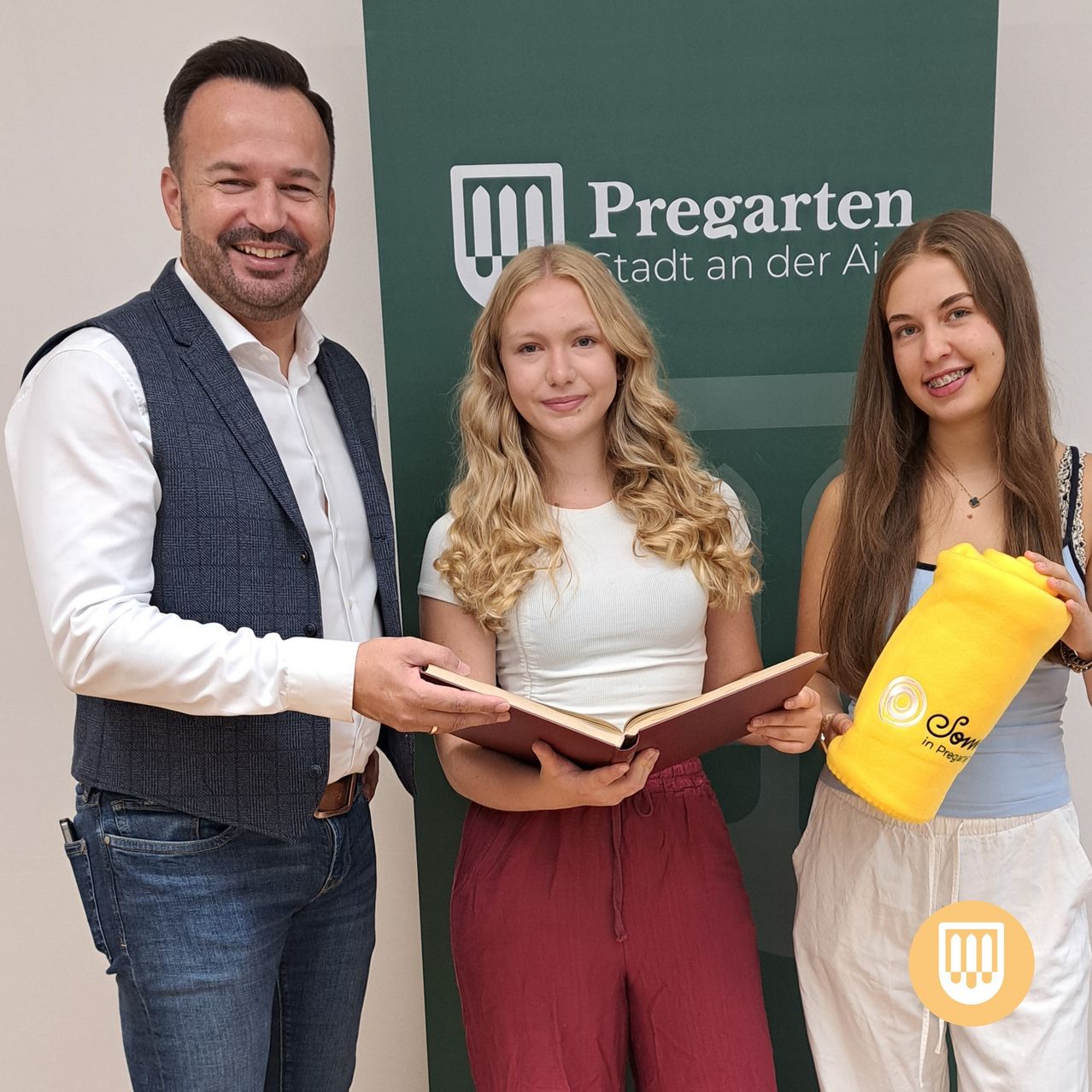 Ein Mann und zwei junge Frauen stehen vor einem grünen Banner mit den Worten 'Pregarten Stadt an der Ai'. Der Mann lächelt und hält ein Buch, während die beiden Frauen lächeln und ein gelbes Objekt halten.