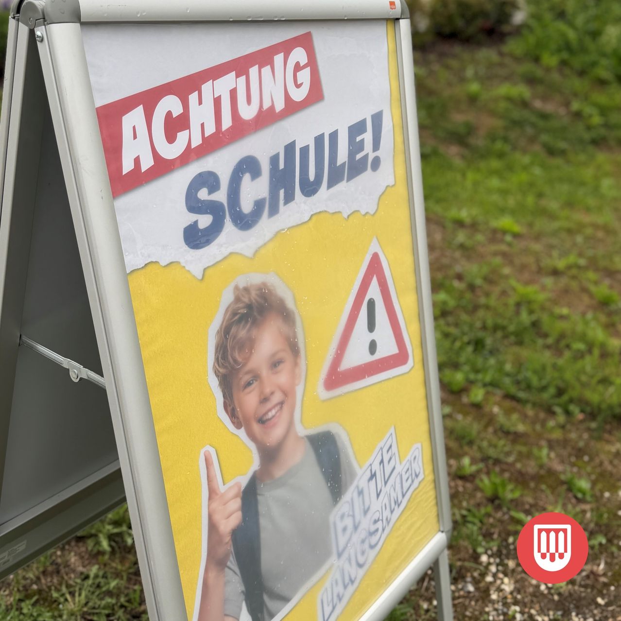 Ein gelbes Schild mit einem Bild eines Jungen und den Worten 'ACHTUNG SCHUL!' in fetten roten Buchstaben. Der Junge gibt einen Daumen hoch und trägt einen Rucksack. Das Schild hat auch ein rotes Dreieck mit einem Ausrufezeichen.