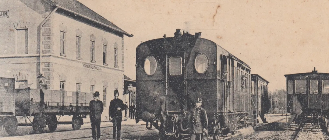 Schwarz-Weiß-Foto eines Zuges auf den Gleisen mit zwei Schaffnern. Das Bild ist beschriftet mit dem Text: 'Gross aus KORNEUBURG - Localbahnhof.'