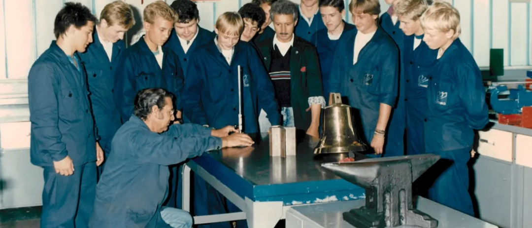Eine Gruppe junger Männer in blauen Uniformen ist um einen Mann versammelt, der kniet und an einem Metallobjekt arbeitet. Er hält ein Werkzeug in der Hand und auf dem Tisch vor ihm befinden sich eine Glocke und ein Amboss. Die Wand hinter ihnen ist mit Postern und einer Topfpflanze dekoriert.