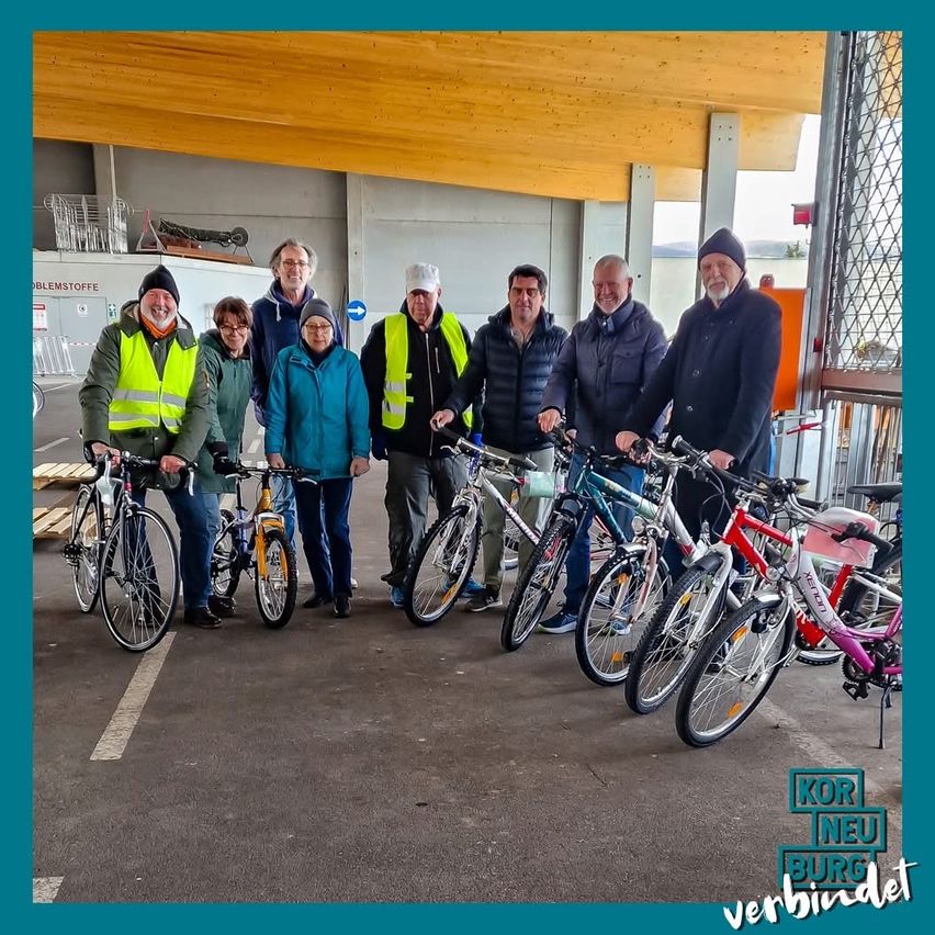 Sieben Personen stehen neben Fahrrädern und tragen Warnwesten und Winterkleidung. Im Hintergrund ist ein Betongebäude mit Holzdach zu sehen.