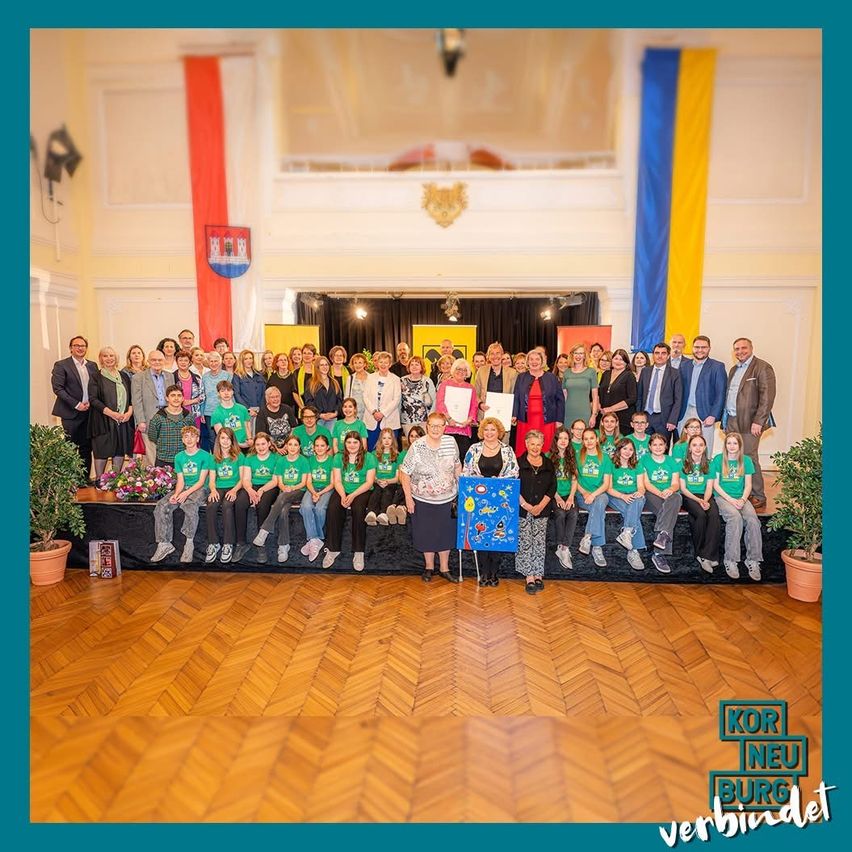 Eine Gruppe von Menschen in grünen Shirts und formeller Kleidung posiert für ein Foto in einem Gebäude mit Holzböden, Fahnen und einer Bühne.