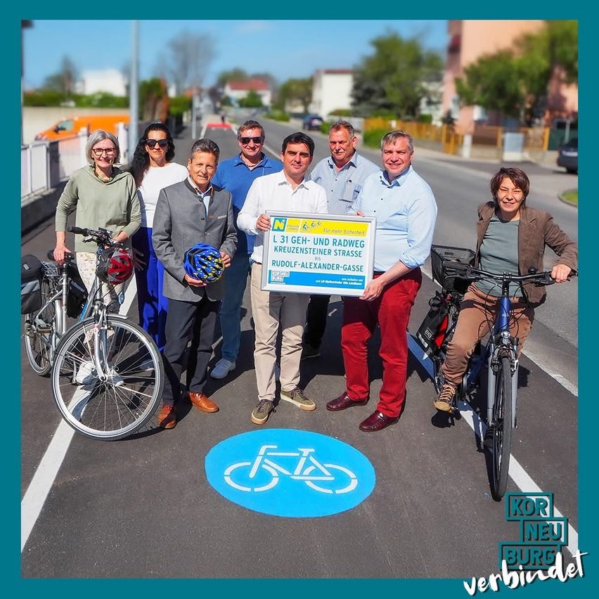 Eine Gruppe von Menschen steht auf einer Straße mit Fahrrädern. Sie halten ein Schild, das die Fertigstellung eines Fahrradwegs anzeigt. Zwei Personen sitzen auf Fahrrädern, während andere Helme halten. Die Straße hat einen neu lackierten Fahrradstreifen.