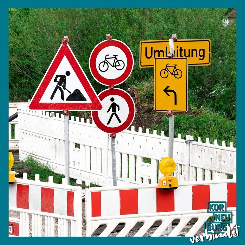 Baustellenschilder und ein gelbes Fahrradumleitungsschild stehen in der Nähe eines weißen Zauns. Im Hintergrund sind Bäume und Grünpflanzen zu sehen.