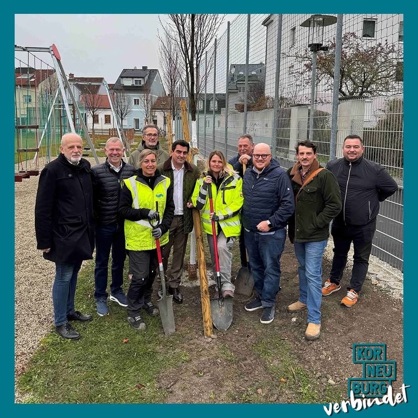 Eine Gruppe von Personen, einige in Warnwesten, posiert mit Schaufeln hinter einem Zaun, mit Gebäuden und einem Spielplatz im Hintergrund.