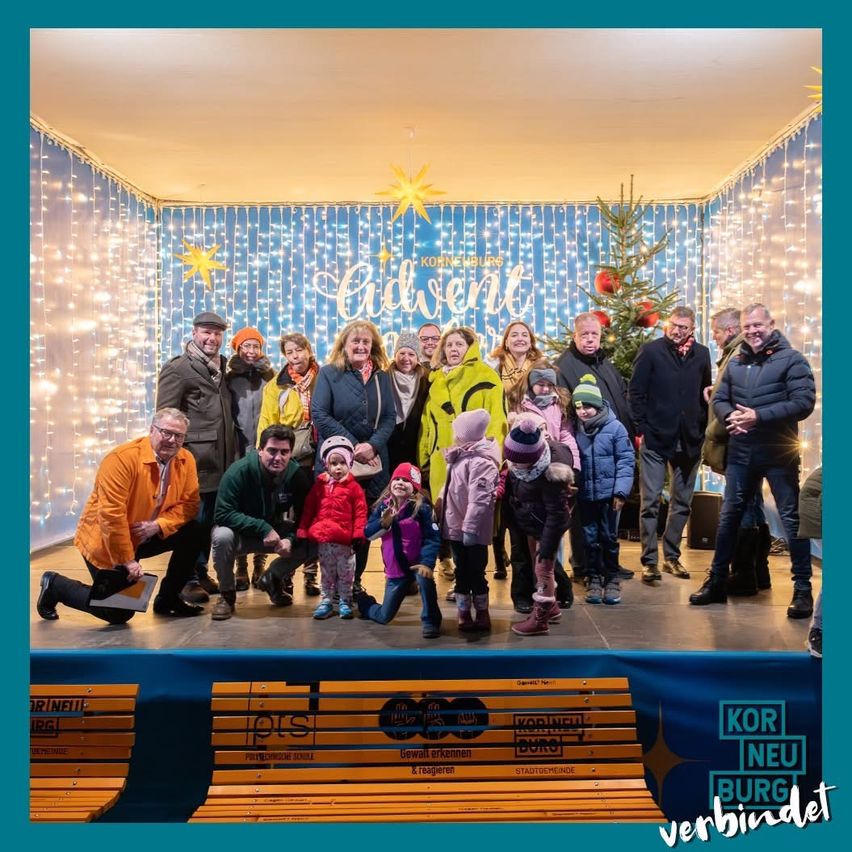 Eine Gruppe von Menschen posiert für ein Foto in einem festlichen Ambiente. Sie sind von Weihnachtsdekorationen umgeben, darunter Lichter und ein Baum. Der Hintergrund zeigt 'Advent.' Die Gruppe besteht aus Erwachsenen und Kindern, alle warm gekleidet für den Anlass. Das Foto wird vor einer Bühne mit einer Holzbank aufgenommen.