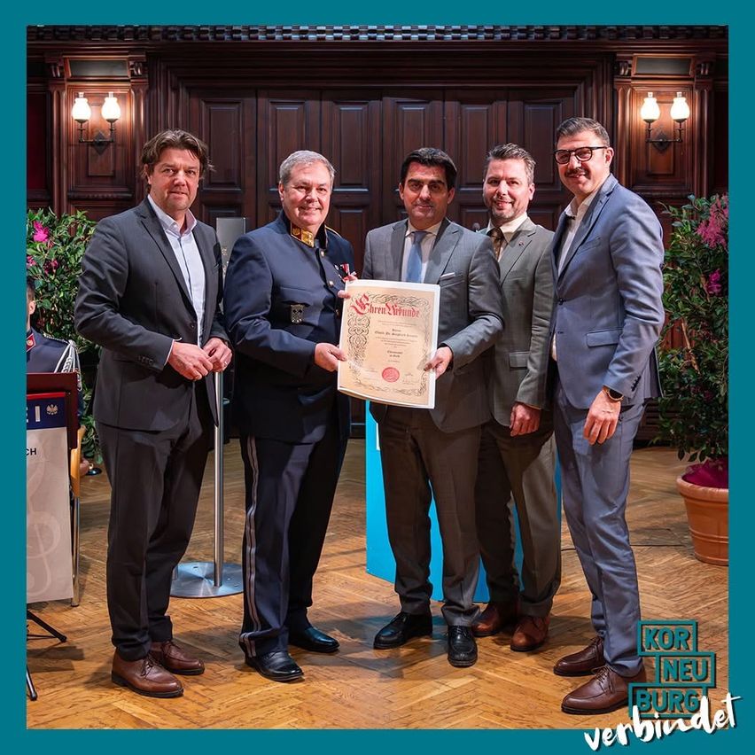 Fünf Männer in Anzügen stehen in einem Raum, einer hält ein Zertifikat. Sie scheinen an einer Preisverleihung teilzunehmen.