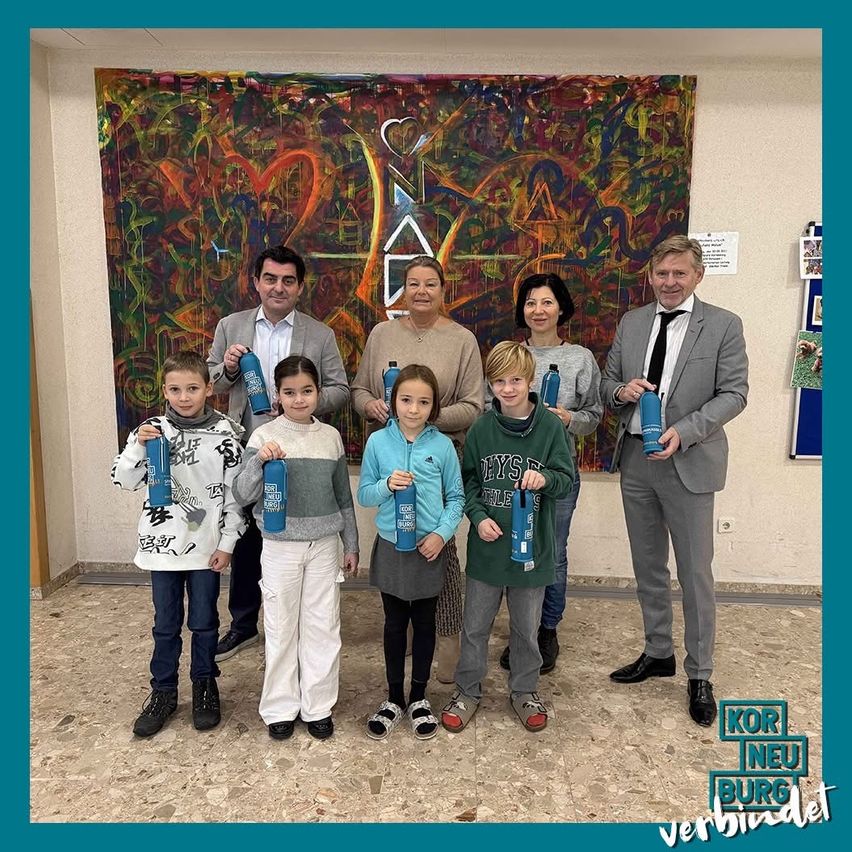 Eine Gruppe von Menschen, Erwachsene und Kinder, steht drinnen vor einem farbenfrohen abstrakten Gemälde und hält blaue Flaschen. Sie lächeln und scheinen für ein Foto zu posieren.