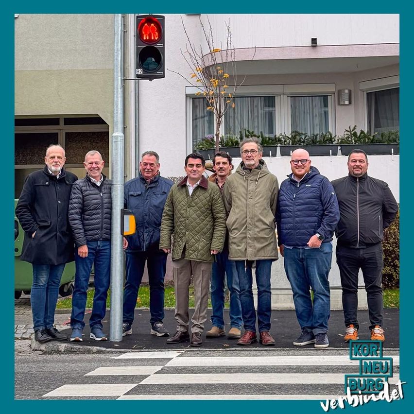 Eine Gruppe von Männern steht auf einem Bürgersteig mit einem Zebrastreifen und lächelt in die Kamera. Sie sind warm gekleidet in Jacken. Dahinter steht eine Ampel an einem Pfosten. Ein Baum und eine kleine Topfpflanze sind in der Nähe. Das Gebäude hat Fenster mit Vorhängen.