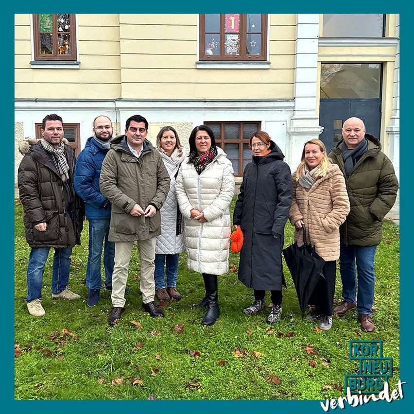 Eine Gruppe von Menschen in Winterkleidung posiert vor einem Gebäude. Das Gebäude hat gelbe Wände und mehrere Fenster. Die Menschen lächeln und scheinen für ein Foto zu posieren.