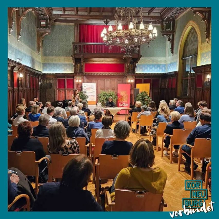 Eine Veranstaltung in einem Raum mit mehreren Menschen, die auf Stühlen sitzen, wahrscheinlich eine Besprechung. Zwei Personen stehen vor einem Tisch, und es gibt Pflanzen und eine Kronleuchter.