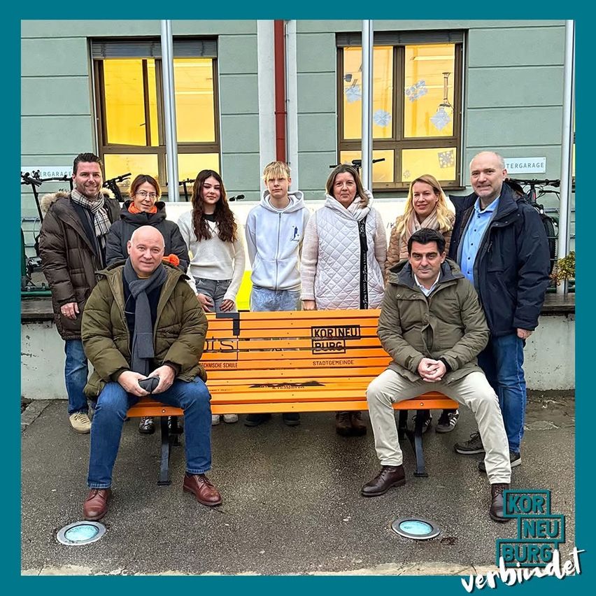 Eine Gruppe von Menschen sitzt und steht hinter einer Holzbank mit der Aufschrift 'Kor Neu Burg' vor einem Gebäude mit gelben Fenstern.