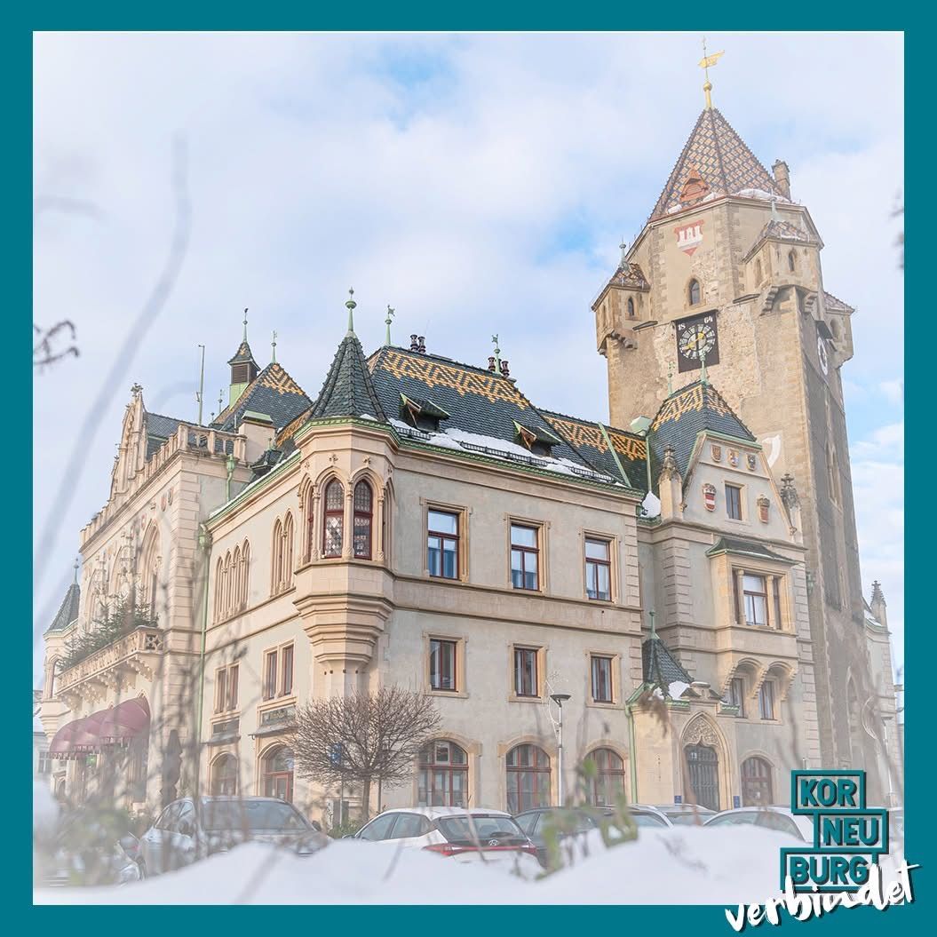 Ein großes schlossartiges Gebäude mit mehreren Türmen und Spitzen. Es hat einen Uhrturm und ist von Autos auf einem verschneiten Parkplatz umgeben.