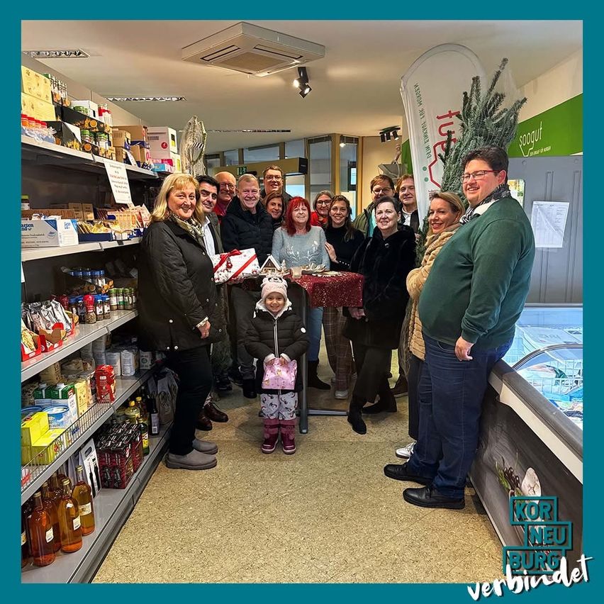 Eine Gruppe von Menschen, darunter Erwachsene und Kinder, versammelt sich in einem Geschäft. Sie stehen vor Regalen, die mit verschiedenen Produkten gefüllt sind, während eine Person ein Geschenk hält und ein Kind einen Hut trägt.