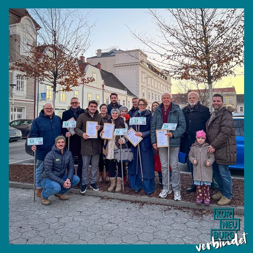 Eine Gruppe von Erwachsenen und Kindern, die in einer Reihe stehen und Urkunden halten. Sie lächeln und posieren für ein Foto. Sie befinden sich in einer Stadt mit Gebäuden im Hintergrund.