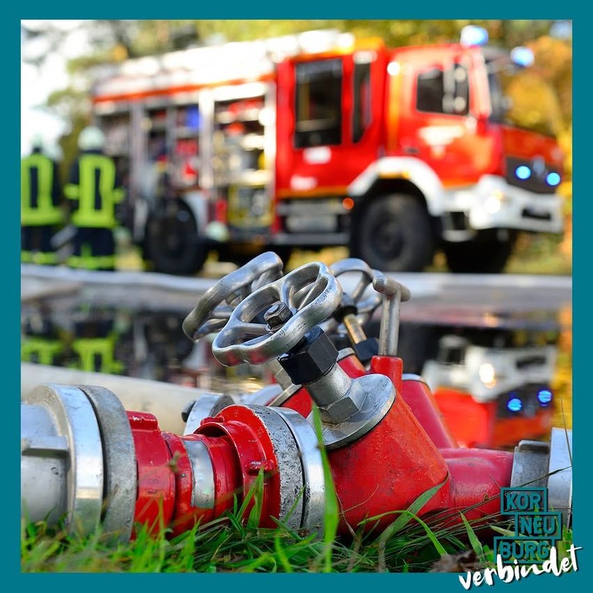 Ein roter Feuerwehrwagen steht am Straßenrand, zwei Feuerwehrleute stehen in der Nähe. Ein roter Feuerhydrant ist im Vordergrund.