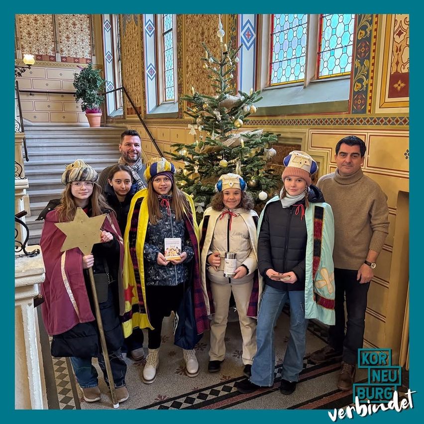 Eine Gruppe von Menschen in Winterkleidung steht neben einem Weihnachtsbaum und lächelt für ein Foto. Dahinter befindet sich eine Treppe mit einer Topfpflanze.