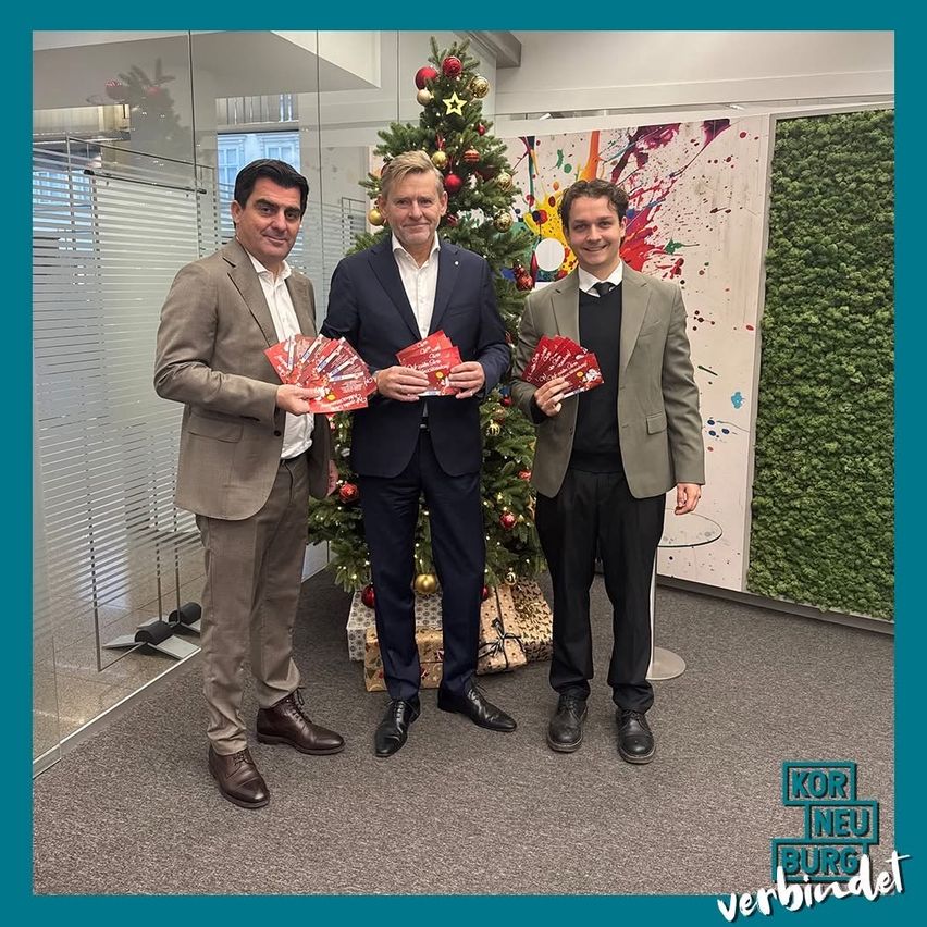 Drei Männer stehen vor einem Weihnachtsbaum und halten rote Karten in der Hand. Sie lächeln und sind in Anzügen gekleidet. Im Hintergrund befindet sich eine Glaswand und ein Weihnachtsbaum.