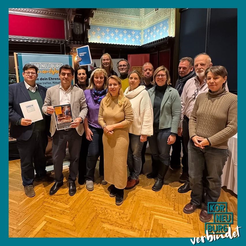 Eine Gruppe von Menschen steht in einem Saal, lächelt und hält ein Buch und eine Broschüre. Hinter ihnen steht ein Banner mit der Aufschrift 'Kor Neu Burg'. Sie sind in Winterkleidung gekleidet, und der Boden ist aus Holz.