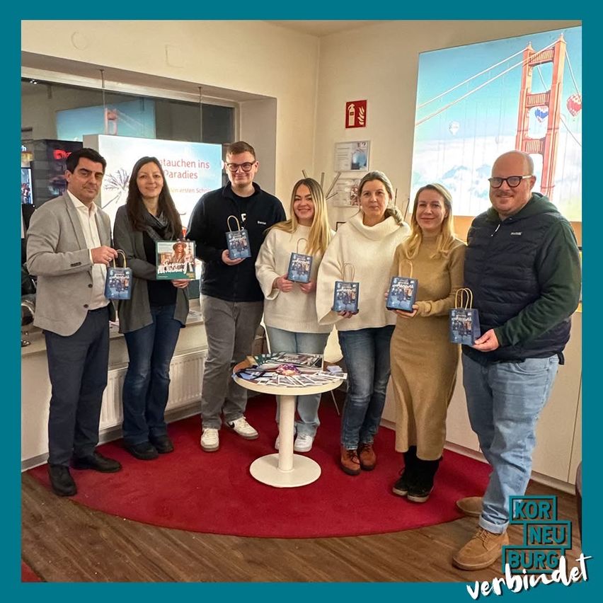 Eine Gruppe von Menschen steht und hält Taschen mit Büchern darin. Sie sind in einem Raum mit einem Tisch und einem Teppich auf dem Boden. Hinter ihnen ist ein Poster einer Brücke und Ballons.