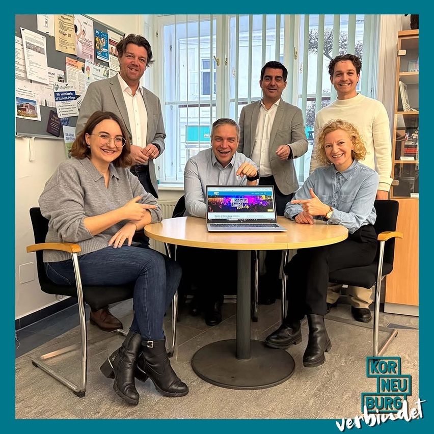 Sechs Menschen sitzen um einen Tisch in einem Raum und lächeln. Ein Laptop steht auf dem Tisch. Zwei Frauen und vier Männer sind anwesend. Ein blauer Rand ist an den Seiten des Bildes.