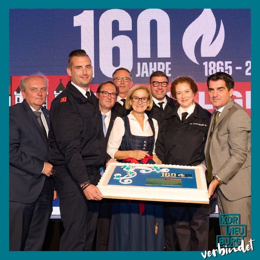 Eine Gruppe von Menschen steht vor einem blauen Hintergrund mit '16n' und 'Jahre' darauf geschrieben. Sie halten einen Kuchen, auf dem '16n' steht. Die Menschen tragen formelle Kleidung.