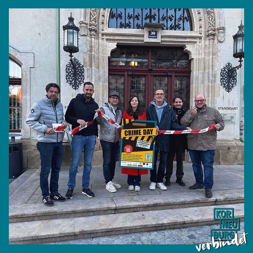 Eine Gruppe von Personen steht vor einem Gebäude, hält ein Banner mit der Aufschrift 'CRIME DAY' und schneidet ein Band durch. Sie sind in Winterkleidung gekleidet und scheinen für ein Foto zu posieren.