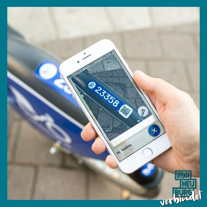 Eine Person hält ein Smartphone mit einer Nahaufnahme eines blauen Fahrradkennzeichens 23358. Der Bildschirm zeigt eine App-Oberfläche mit einer Taschenlampen-Symbol und einem QR-Code. Das Fahrrad steht auf einem Ziegelstein-Gehweg geparkt.