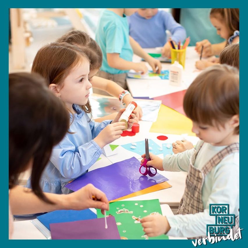 Mehrere junge Kinder sind in einer Bastelaktivität verstrickt, um einen Tisch herum sitzend. Jedes Kind hat eine Schere und Papier vor sich. Die Kinder tragen blaue Hemden. Ein grünes und weißes Banner in der unteren rechten Ecke lautet 'KOR NEU BURG verbindet'.