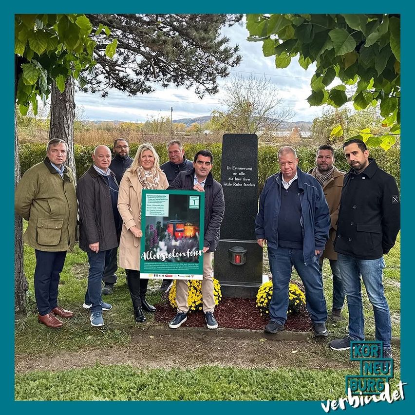 Eine Gruppe von Menschen steht in einem Park, einige halten ein grünes Schild und andere in der Nähe eines Denkmals. Das Denkmal trägt die Aufschrift 'In Erinnerung an alle, die hier ihre letzte Ruhe fanden'. Das Bild hat einen Wasserzeichen, der 'Korneuburg verbindet' lautet.