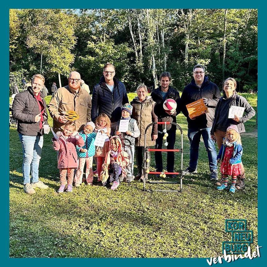 Eine Gruppe von Menschen posiert für ein Foto in einem Park mit Bäumen im Hintergrund. Einige halten Urkunden und Sportgeräte, während ein Kind mit einer kleinen roten Leiter spielt.