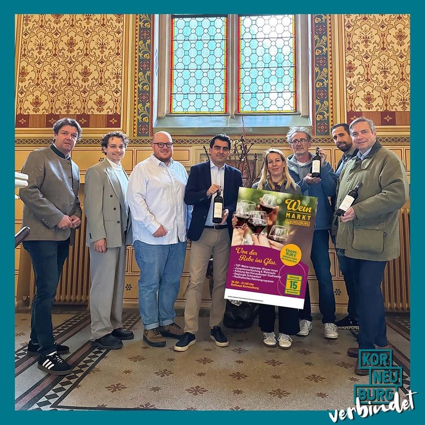 Eine Gruppe von Menschen steht drinnen, hält Weinflaschen und Gläser und posiert wahrscheinlich für ein Foto. Hinter ihnen befindet sich ein großes Fenster mit dekorativen Mustern.