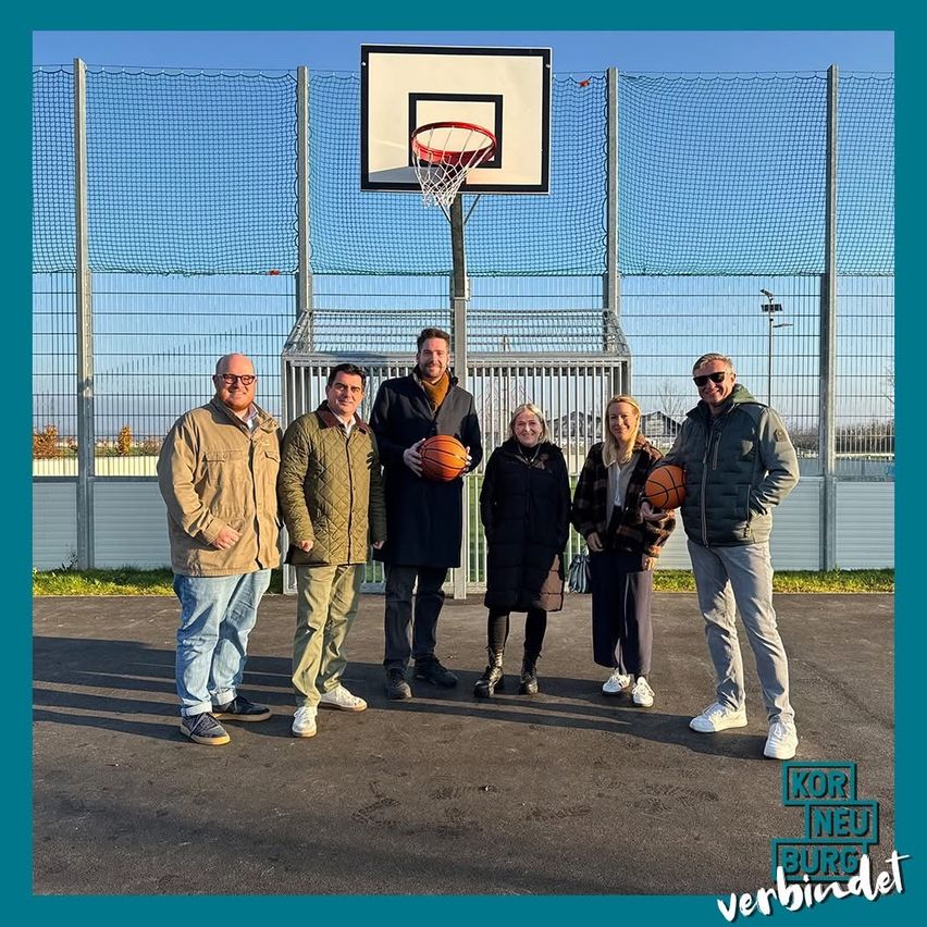 Fünf Personen stehen vor einem Basketballplatz. Drei Männer und zwei Frauen, alle in Winterkleidung, zwei Männer halten Basketballs.