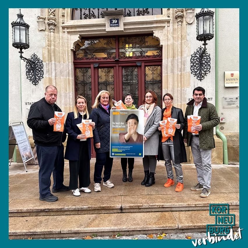 Eine Gruppe von Menschen steht auf den Stufen eines Gebäudes und hält ein orangefarbenes Plakat mit Text und einem Bild einer Frau. Sie sind draußen.
