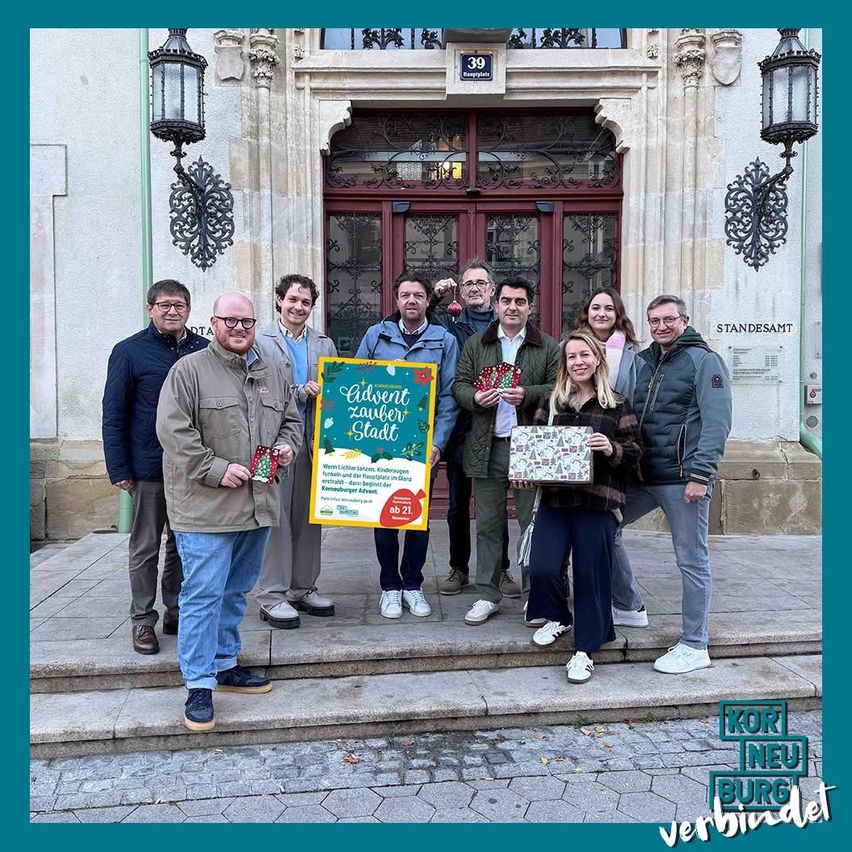 Eine Gruppe von Menschen steht vor einem Gebäude, lächelt und posiert für ein Foto. Sie halten ein Schild mit der Aufschrift Advent Zaubert Stadt und tragen Winterkleidung.
