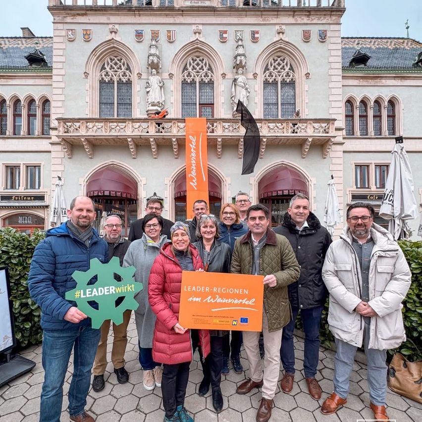 A group of people, some wearing glasses, stand in front of a historic building. They hold signs and appear to be posing for a photo.