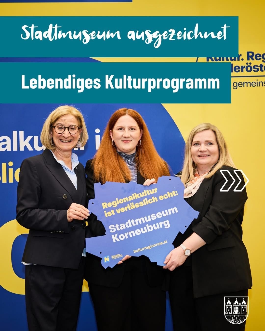 Three women stand together holding a blue placard with a yellow outline. They are in front of a yellow and blue backdrop. The text on the placard says Regionalkultur ist verlasslich echt.