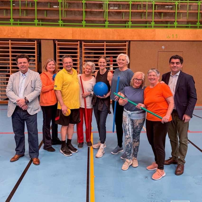 A group of people are standing in a sports hall, smiling and posing for a photo. They are holding rackets and a ball. Behind them is a wooden wall and an open door.
