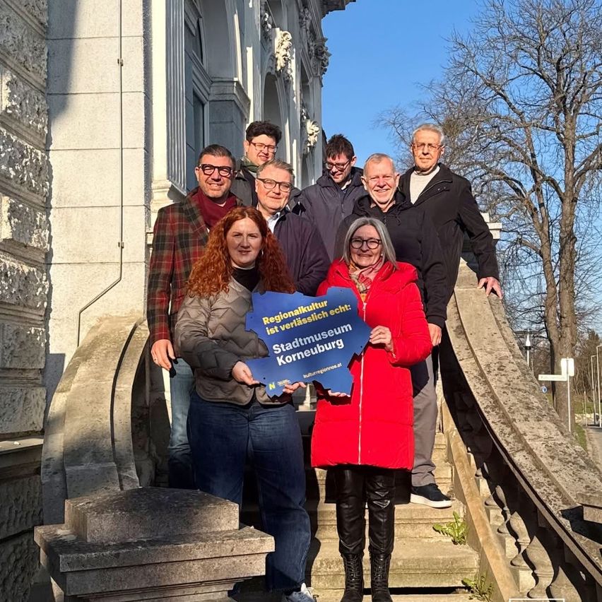 A group of people, both men and women, are standing on the stairs of a building. They are smiling and appear to be posing for a photo. A woman is holding a blue sign with white text.