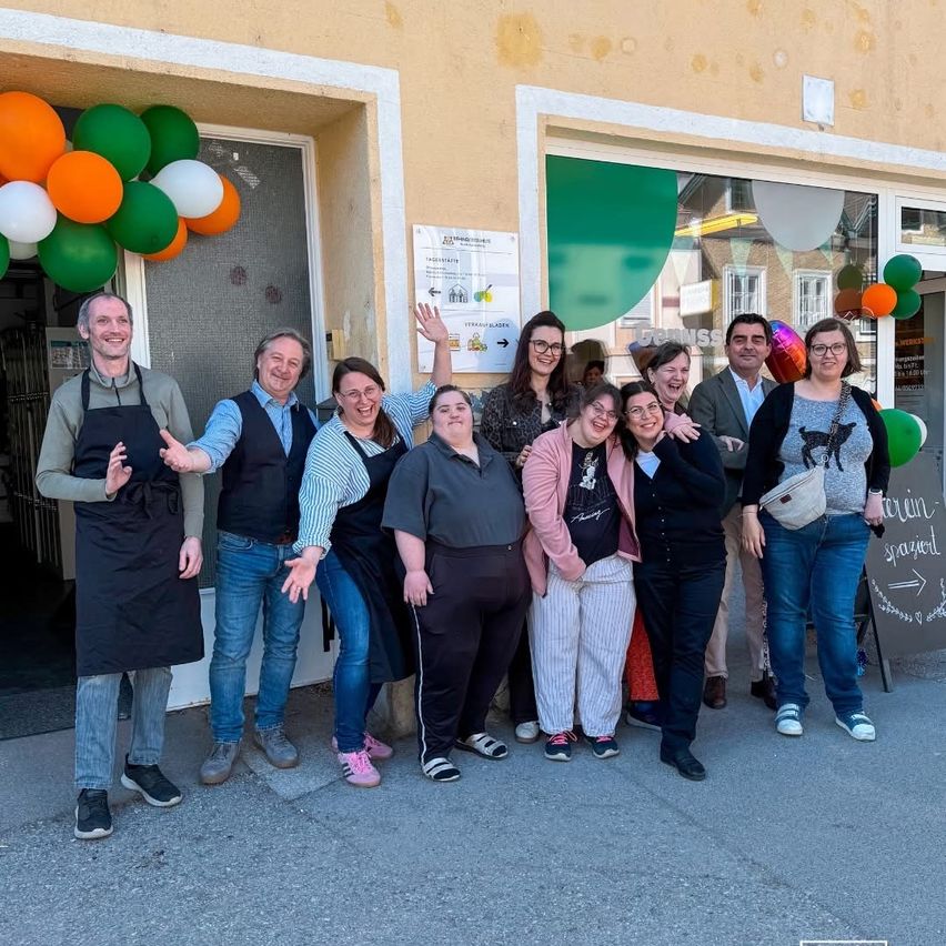A group of people, dressed casually, stand outside a shop. They are posing for a photo with balloons in the background. Some are wearing glasses and aprons.