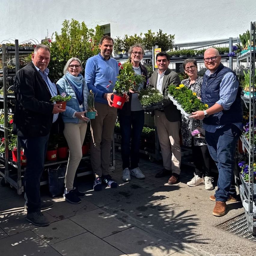 A group of seven people are standing outside, posing for a photo. They hold various potted plants. The background shows a garden with many more plants and flowers.