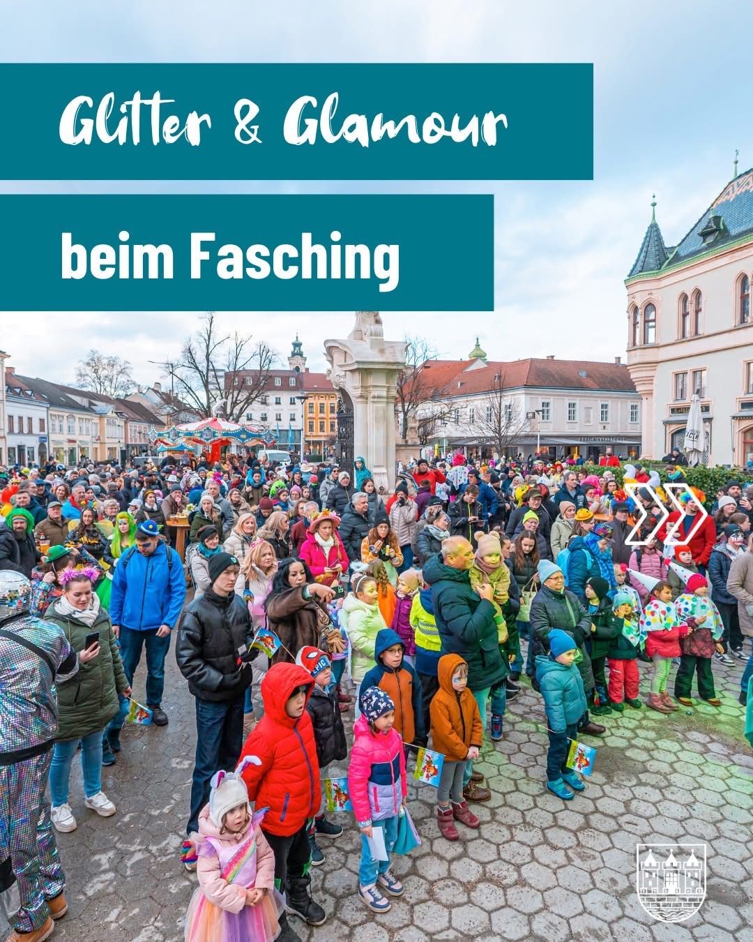 A crowd gathers in a town square for Fasching, a festive celebration with many people dressed in colorful costumes, including children holding flags and adults with painted faces, under a cloudy sky.