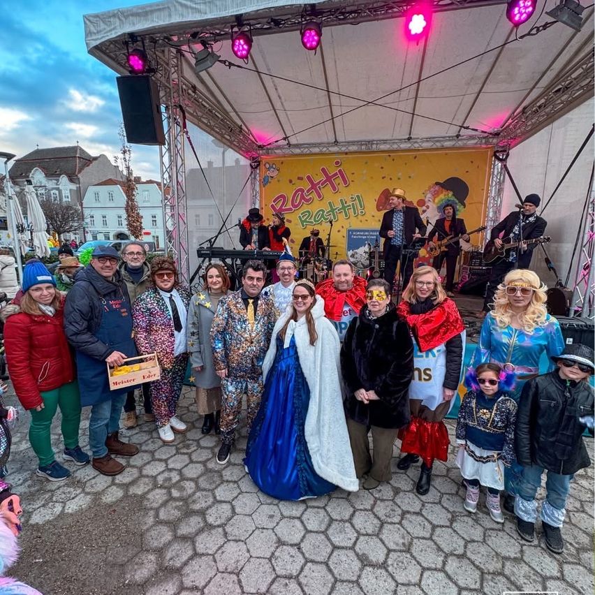 A group of people dressed in various costumes pose for a photo on a stage with a banner reading 'Ratti Ratti!' behind them.