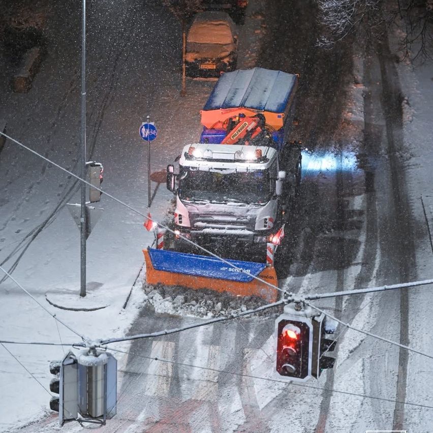 A snowplow truck is clearing snow from the road at night. The road is covered with snow, and there are traffic lights and utility poles on either side.