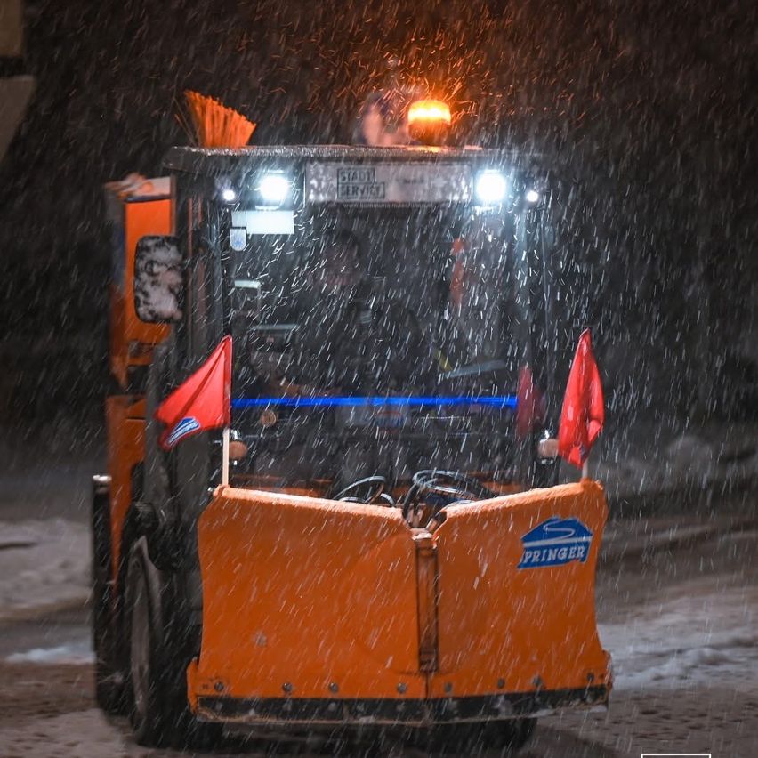 Ein Schneepflug mit Springer-Logo auf der Front räumt bei starkem Regen die Straße in der Nacht. Zwei rote Flaggen sind an der Vorderseite des Fahrzeugs befestigt.
