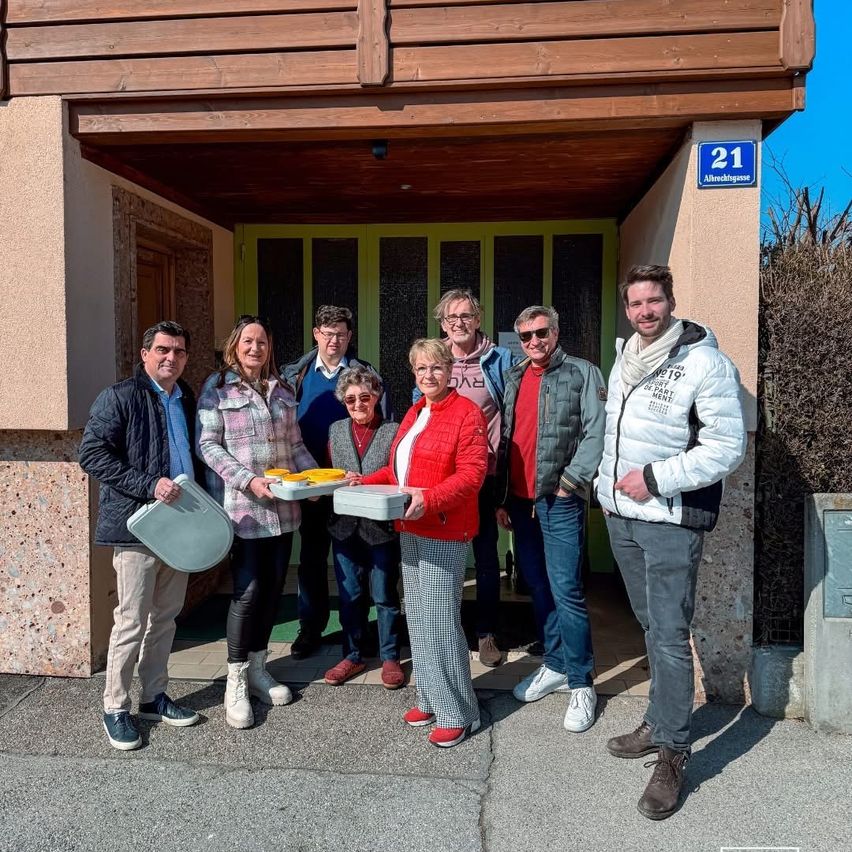 Eine Gruppe von Menschen steht vor einem Gebäude mit der Nummer 21 an der Wand, lächelt und hält Tabletts mit Essen in den Händen. Sie sind in Winterkleidung gekleidet.