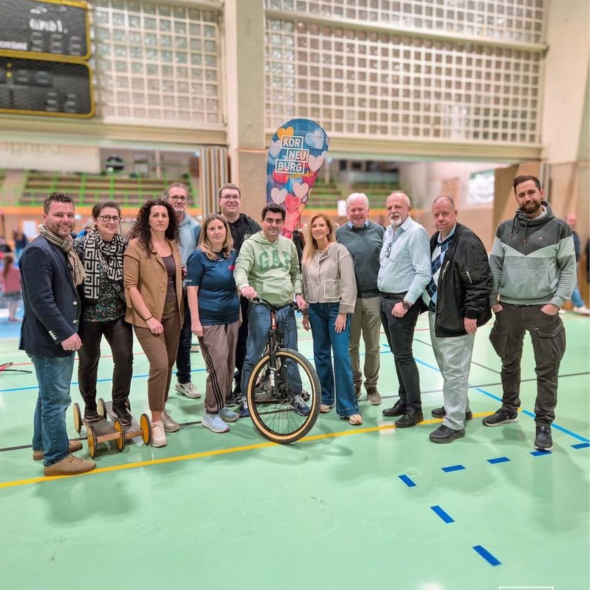 Eine Gruppe von Menschen steht in einer Turnhalle mit einem Banner hinter ihnen, einige tragen Brillen und Turnschuhe. Ein Mann hält ein Fahrrad, und sie scheinen alle für ein Foto zu posieren. Der Boden ist mit blauen Linien markiert.