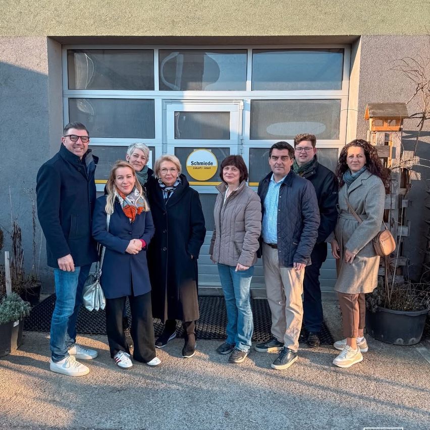 Eine Gruppe von Menschen in Winterkleidung steht vor einem Gebäude mit einem Schild, auf dem 'Schmiede' steht. Sie lächeln und posieren für ein Foto.